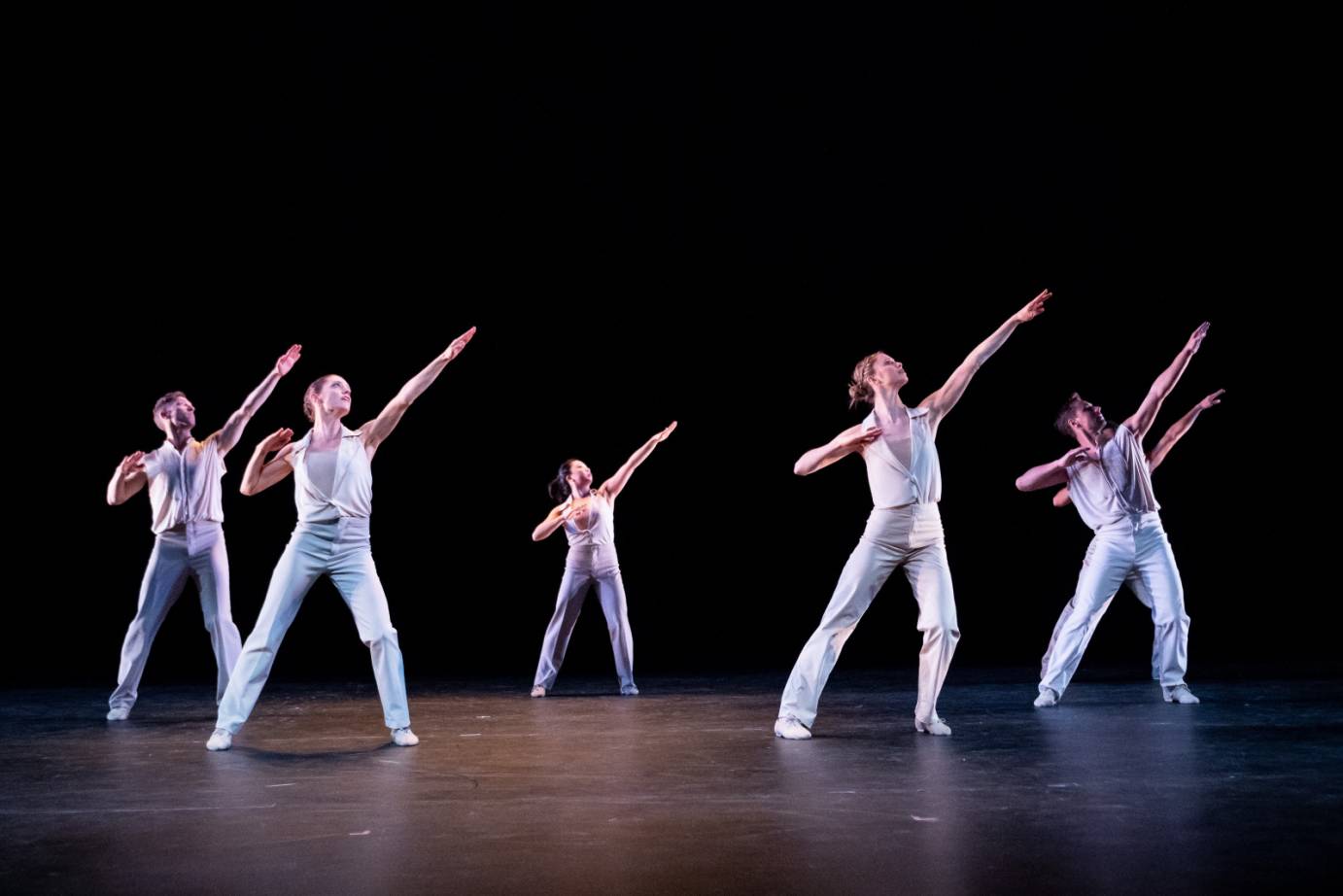 A group of dancers in white pants in tops stand in a wide stance. Their left arm is placed on a high diagonal while their right arm is bent at the elbow.